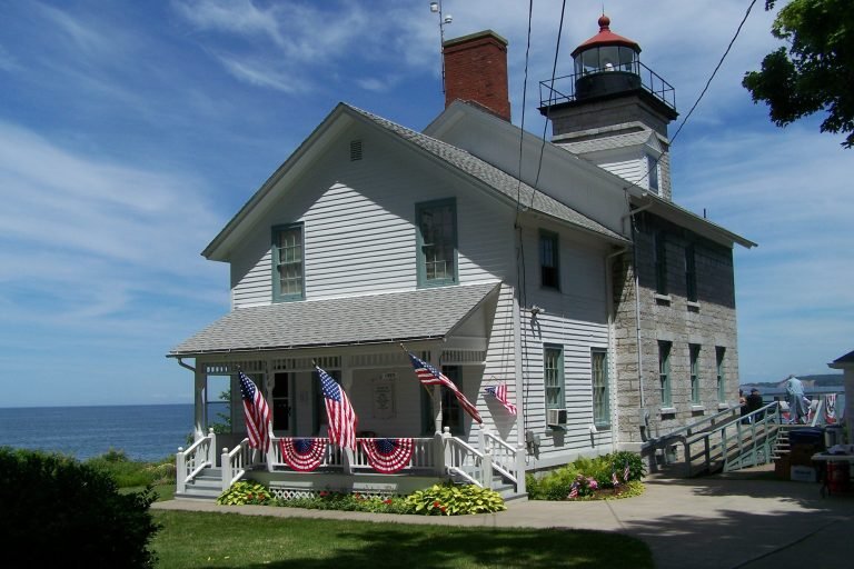 Sodus Bay Lighthouse: white clapboard keeper's house with a flag-draped porch, joined to a stone tower, lake behind