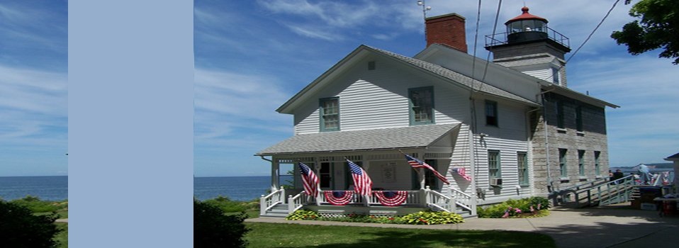 White clapboard house with flag-draped porch joined to a stone lighthouse tower, lake behind, pale blue band at left