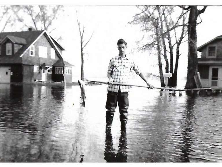 Larry Briggs in waders standing in floodwater between houses, holding a spear with a fish on its end
