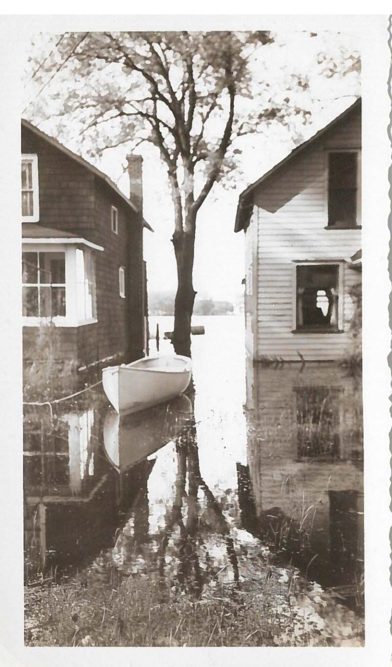 Rowboat floating in floodwater between two houses, a tree standing in the water and the lake beyond