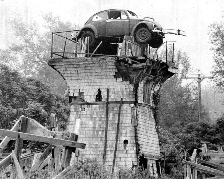Volkswagen Beetle perched atop a crumbling shingled wooden tower, with broken timbers around its base