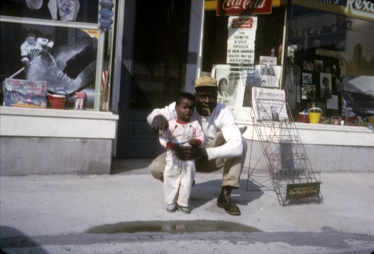 Man in a cap crouching with a small child on the sidewalk outside a store with a Coca-Cola sign