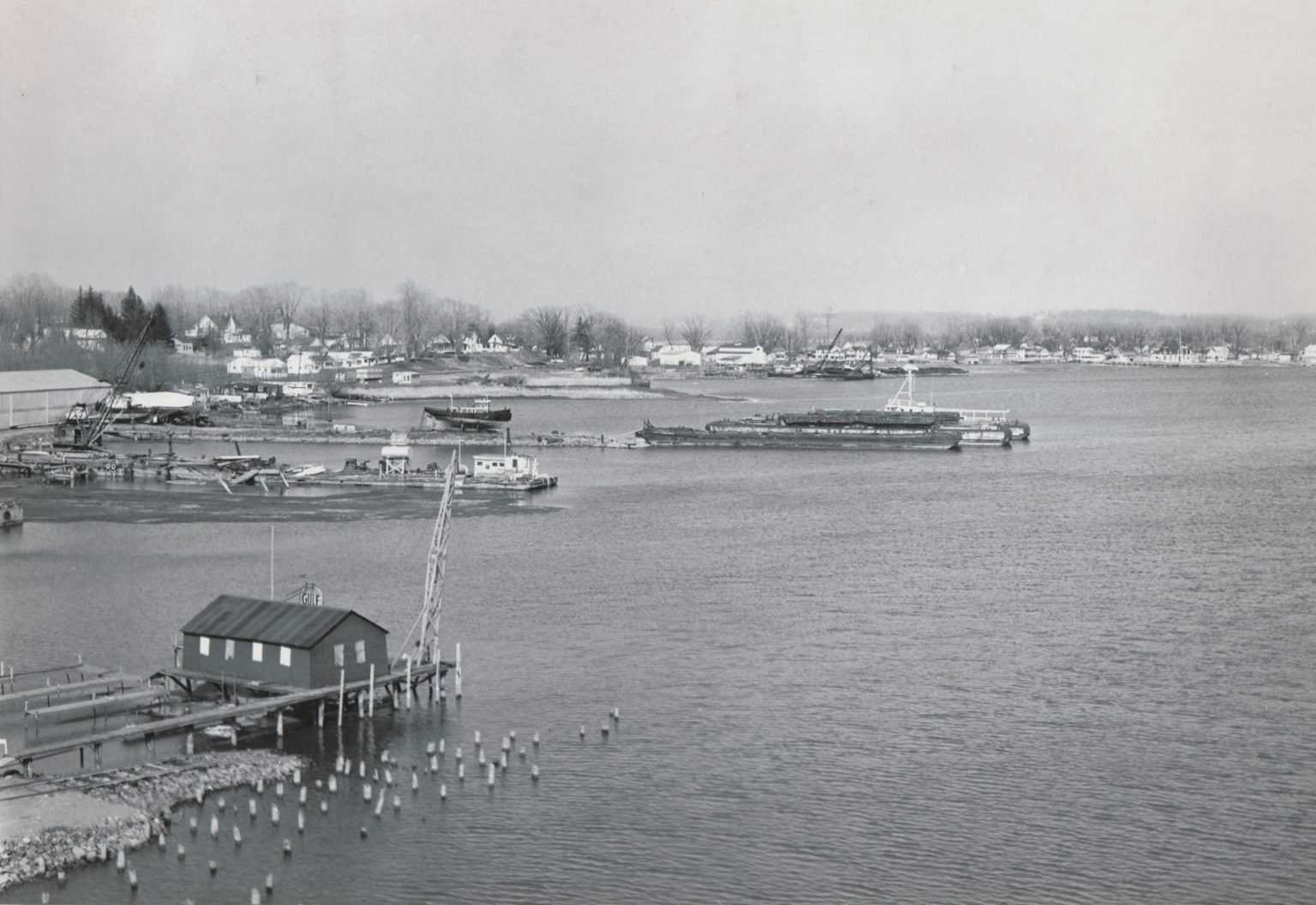 Elevated view across the bay: a boathouse on pilings, barges and a crane, houses along the far shore