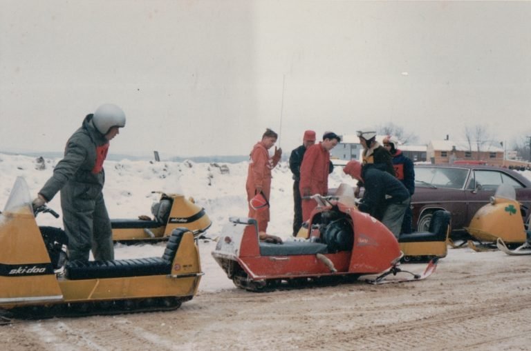 Yellow and orange snowmobiles lined up in the snow, riders in snowsuits and helmets standing beside them