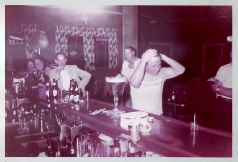 Pink-tinted snapshot of a crowded bar, a man in a white T-shirt with both hands on his head