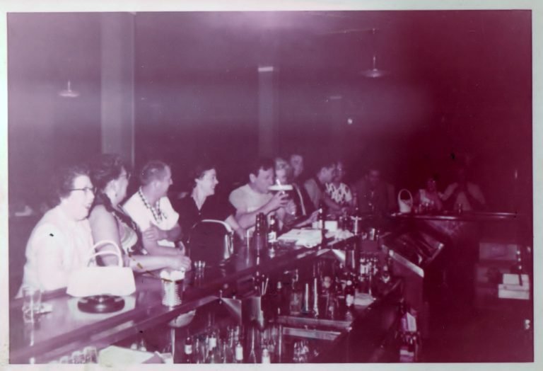 Faded photo of men and women seated along a long bar lined with liquor bottles