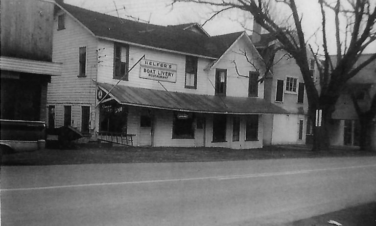 Two-story clapboard building with a 'Helfer's Boat Livery Restaurant' sign above a long porch roof, beside the road