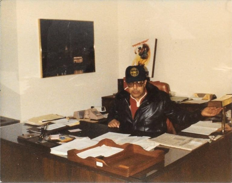 Man in a cap, tinted glasses and leather jacket seated at a large office desk covered with papers