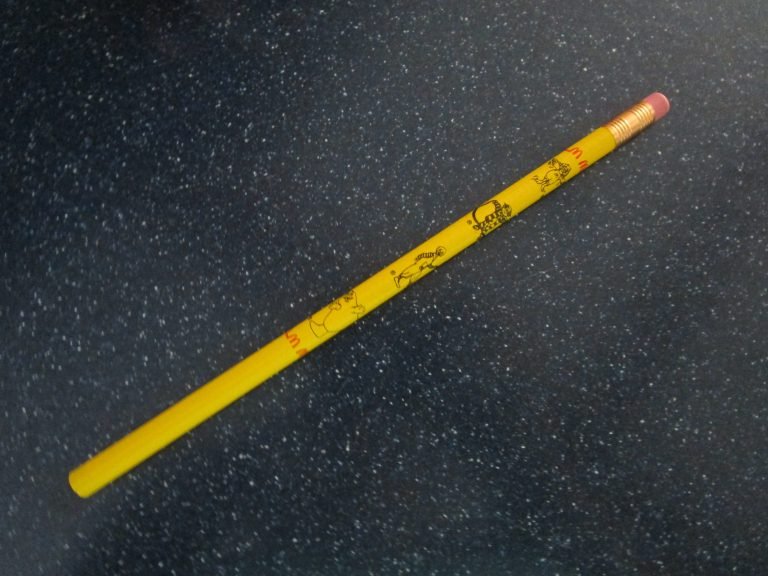 Yellow pencil printed with cartoon characters and red letter M logos, lying on a dark speckled countertop