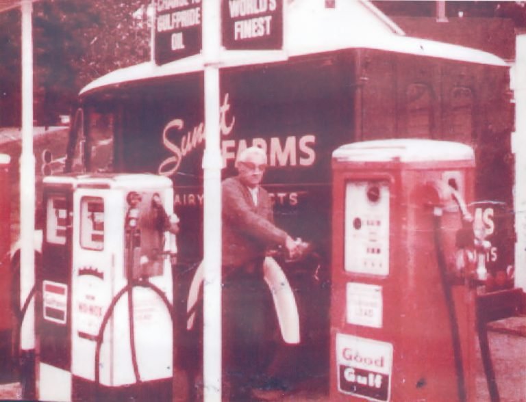 Tal Jones in glasses and a cardigan pumping gas between Gulf pumps, a dairy delivery truck behind