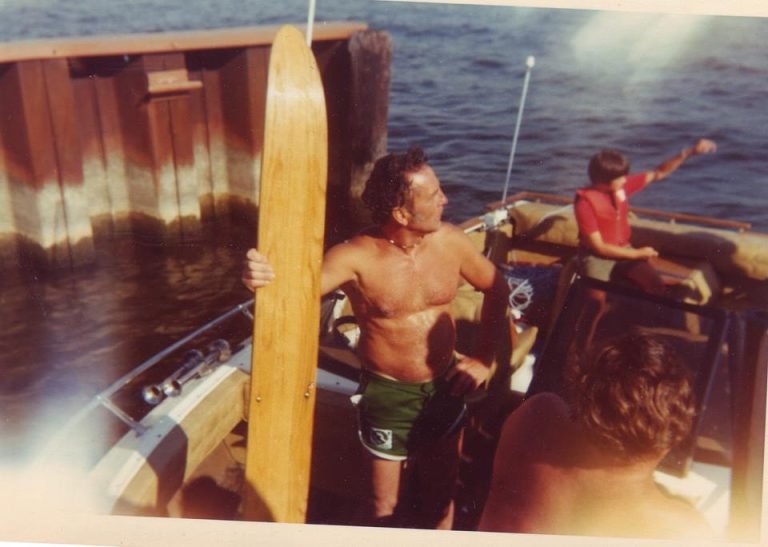 Shirtless Frank Monje holds a wooden water ski aboard a boat beside a pier, friends nearby