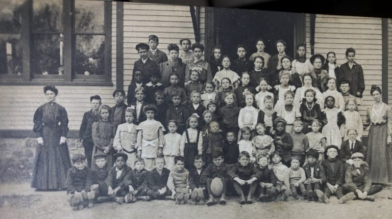 Large group of schoolchildren in rows before a clapboard building, a woman in a long dark dress at each end