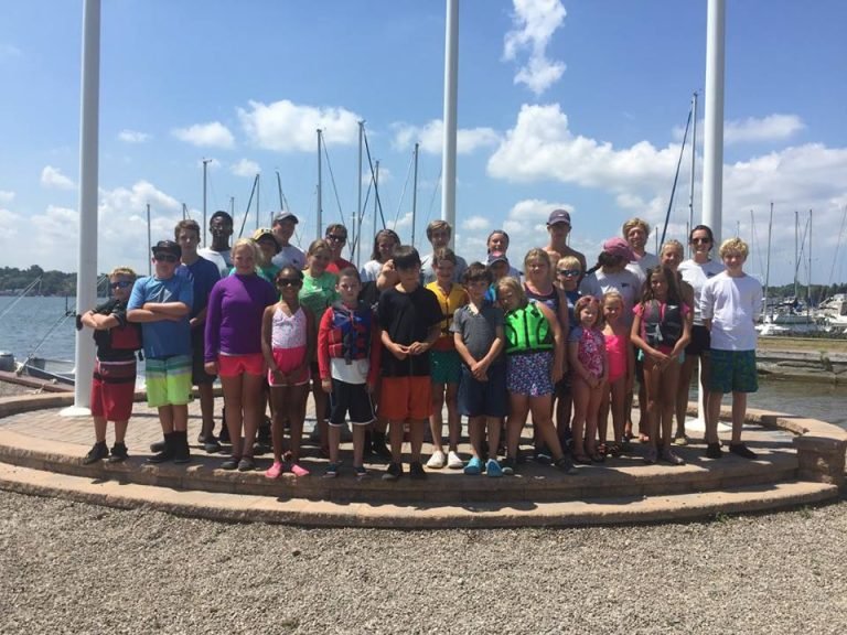 Large group of children and instructors posed by flagpoles at a marina, sailboat masts behind