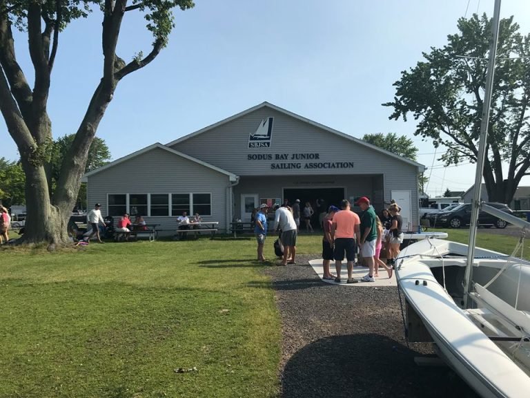 Sodus Bay Junior Sailing Association clubhouse with people gathered on the lawn and a small sailboat in front