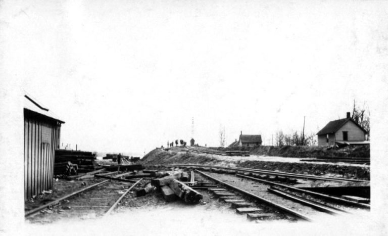 Railroad tracks with piles of heavy timbers, a metal shed at left and small houses on the rise beyond