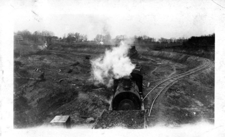 Steam locomotive puffing smoke as it hauls a loaded car along curving tracks through a dug-out cut
