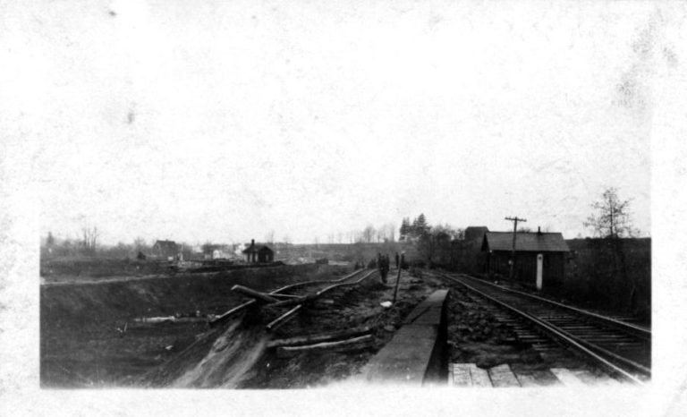 Railroad tracks beside an excavated embankment with scattered timbers, a few workers and small buildings beyond