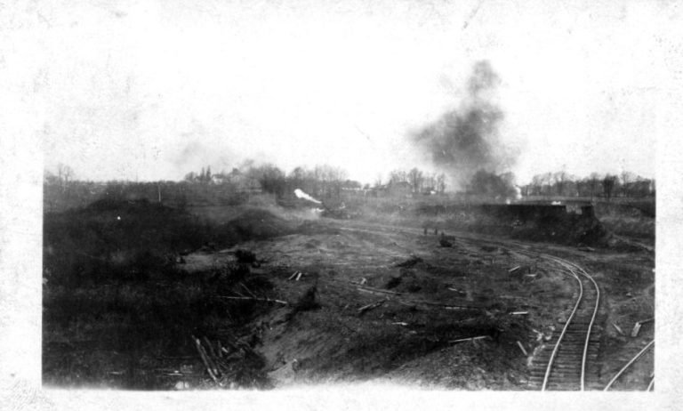 Torn-up muddy ground with a curving rail track, smoke and steam rising in the distance