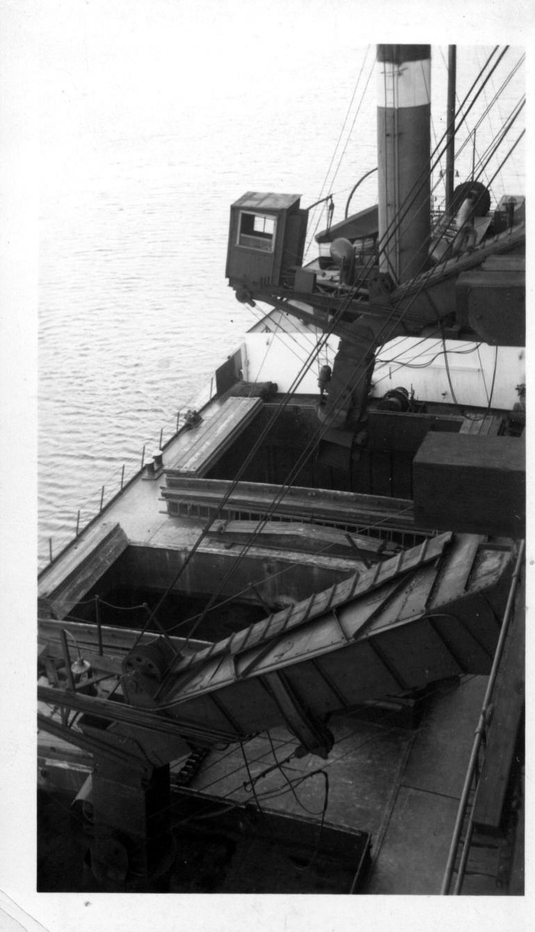 Looking down on a freighter's open hatches as a coal chute and loader cab hang above, the ship's funnel beyond