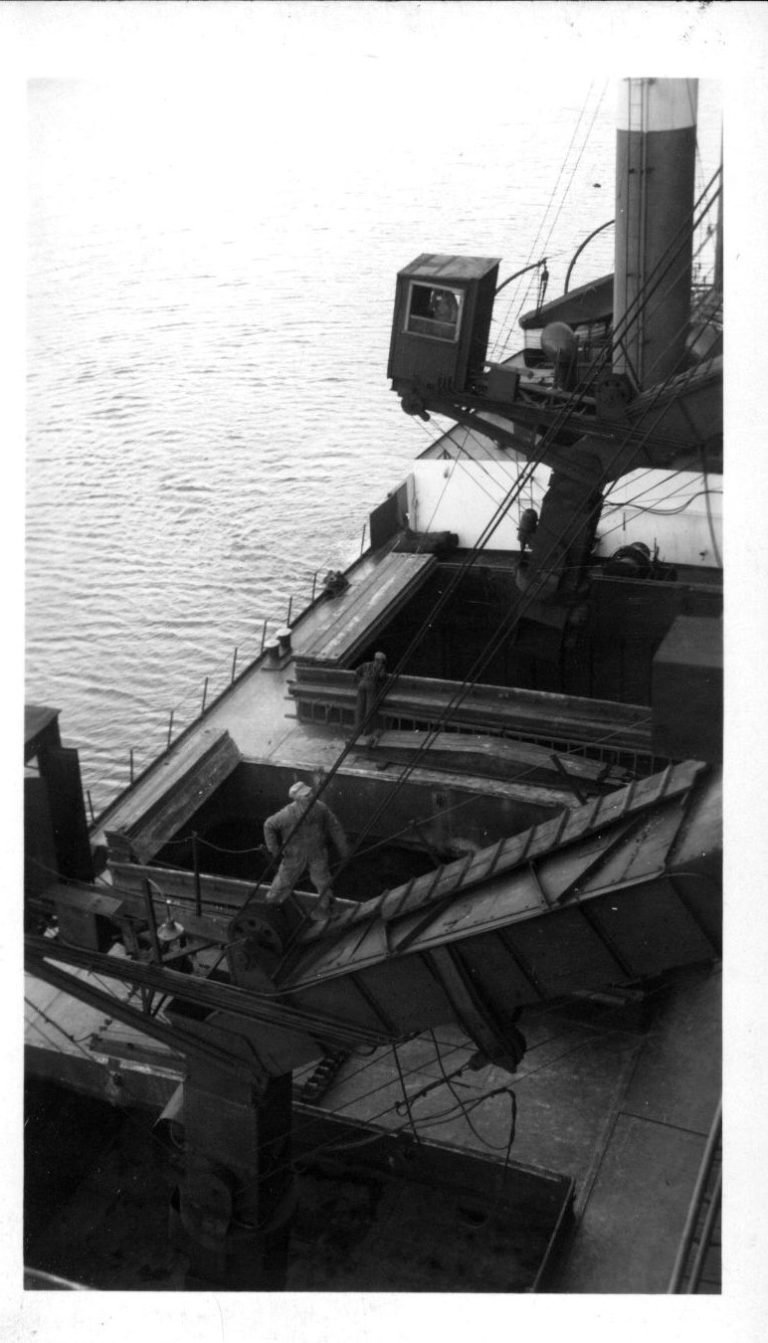 Looking down on a ship's deck as a worker stands by a steel coal chute lowered toward an open hatch