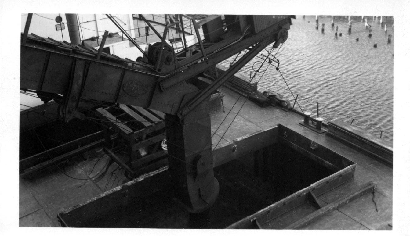 Steel loading arm positioned over a ship's open cargo hatch, pilings in the water beyond