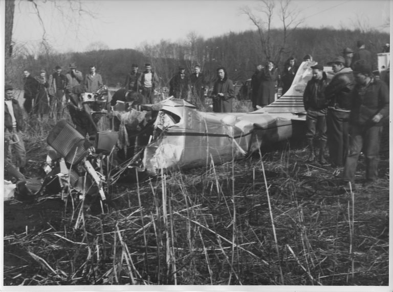 Crowd of men in coats gathered around a small airplane wreck lying in tall marsh grass
