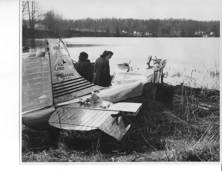 Wrecked small Cessna airplane resting on a grassy shore of the bay, two people looking at it