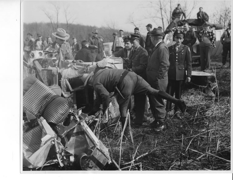 Uniformed officers and onlookers gathered around airplane wreckage in marsh grass, one man bending to examine it