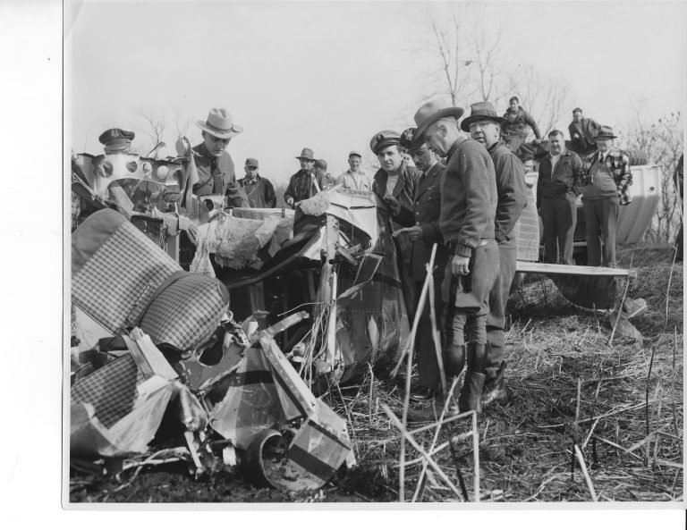 Men in hats and uniformed officers gathered around the crumpled wreckage of a small plane in a brushy field