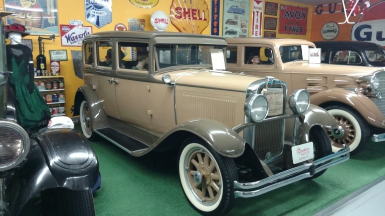 Two tan antique sedans with whitewall tires on display in a room hung with Shell and other old advertising signs