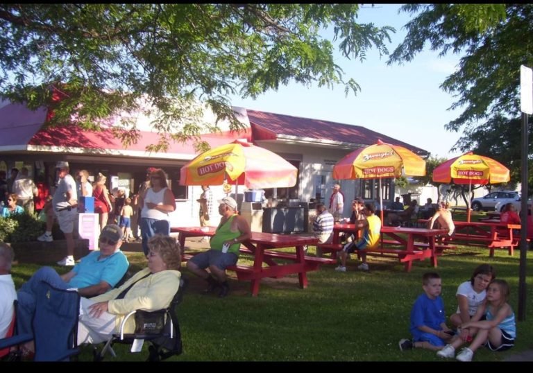 Crowd at red picnic tables under yellow hot dog umbrellas outside Hots Point, a red-roofed snack bar