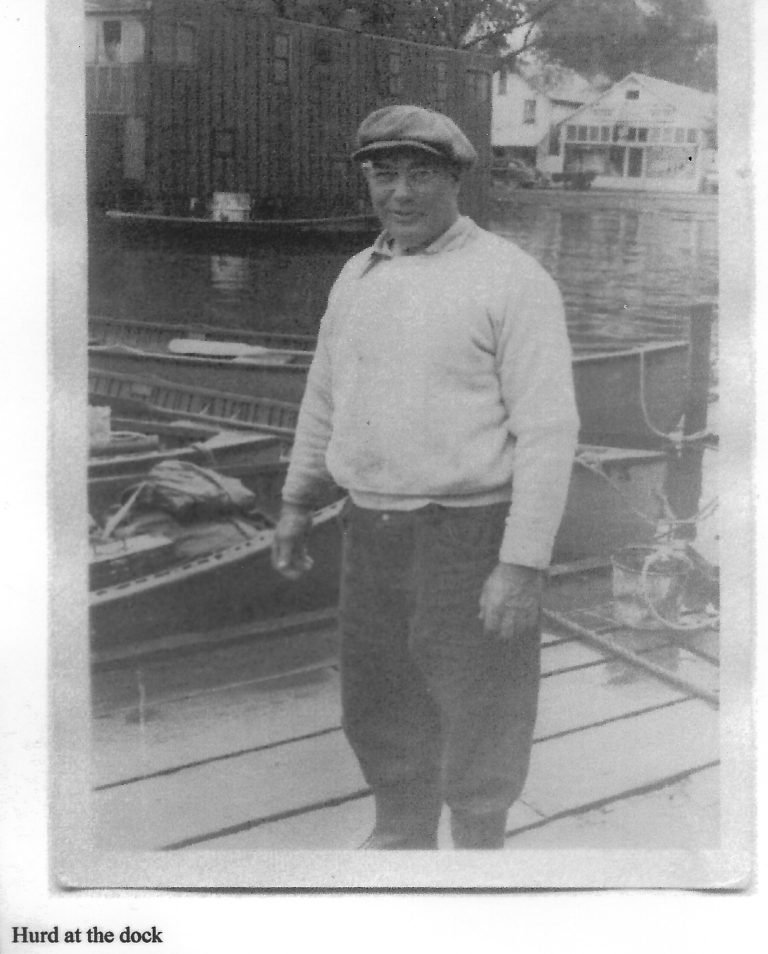 Snapshot captioned 'Hurd at the dock': a man in a flat cap and sweater on a dock by moored boats