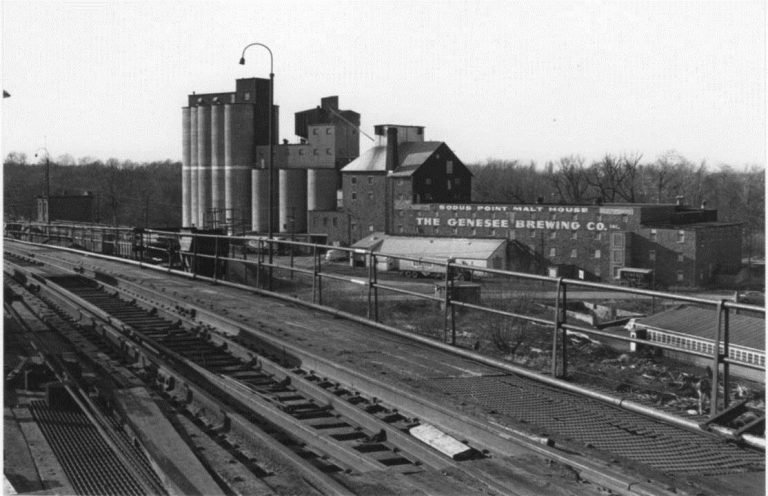 The Malt House, a brick building lettered 'The Genesee Brewing Co.' beside tall silos, seen from the trestle
