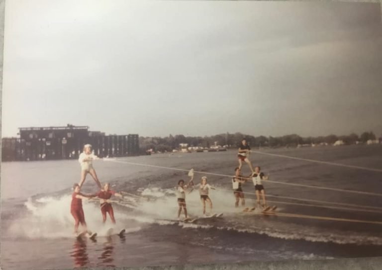 Water skiers forming pyramids while towed across the bay, a large building on the far shore