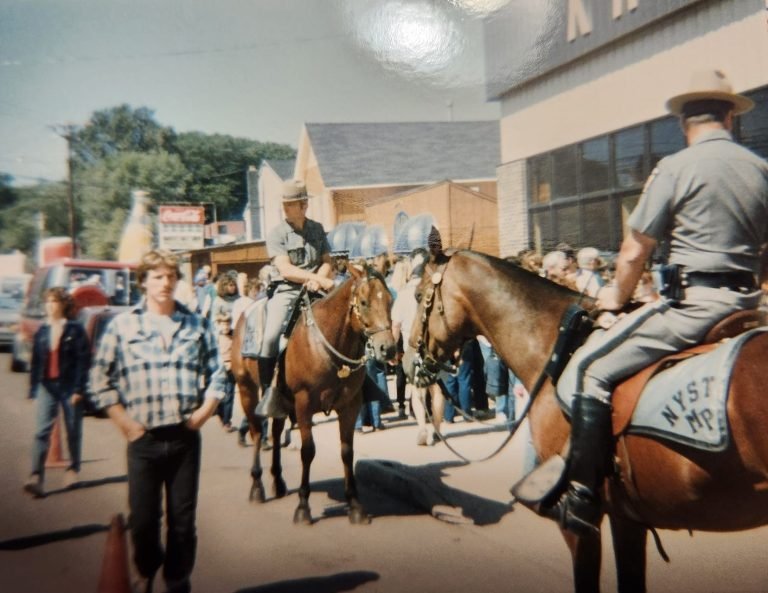 Mounted state troopers in campaign hats on horseback among a crowd on a village street
