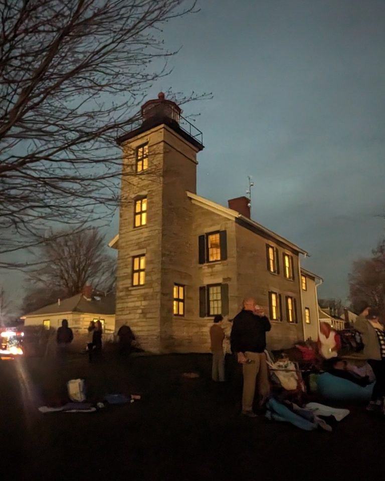 Crowd gathered in the eclipse darkness beside a stone lighthouse and attached house with lit windows
