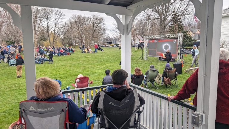 Spectators in camp chairs on a porch and lawn watching a large screen showing the eclipse