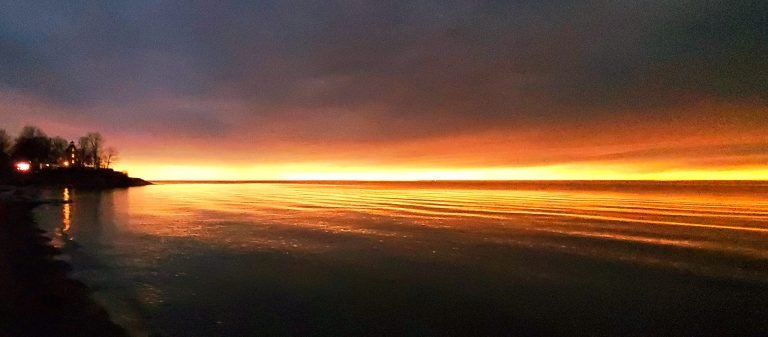 Orange glow along the horizon under dark clouds during the eclipse, trees and buildings silhouetted on a point at left