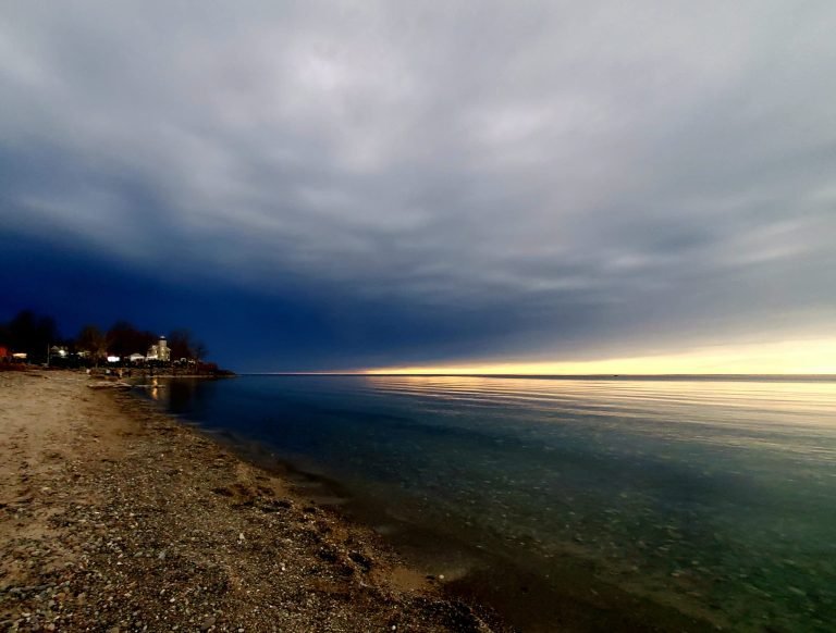 Darkening sky over a calm lake and pebble beach, with a glow on the horizon and buildings along the shore