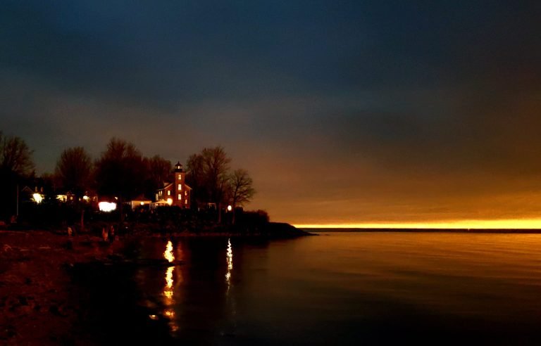Lighthouse with lit windows on the shoreline under a dark sky, an orange glow along the lake horizon