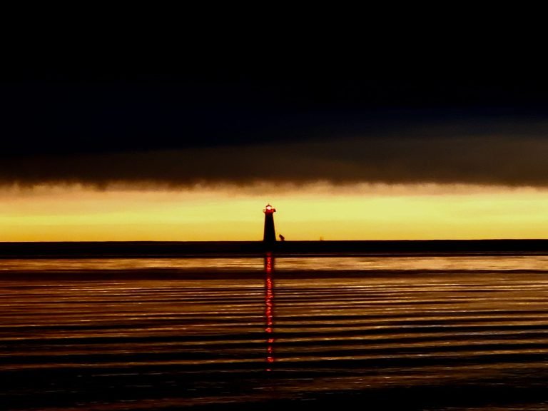 Distant pier lighthouse with its red light reflected across rippled water, under dark clouds and a gold horizon