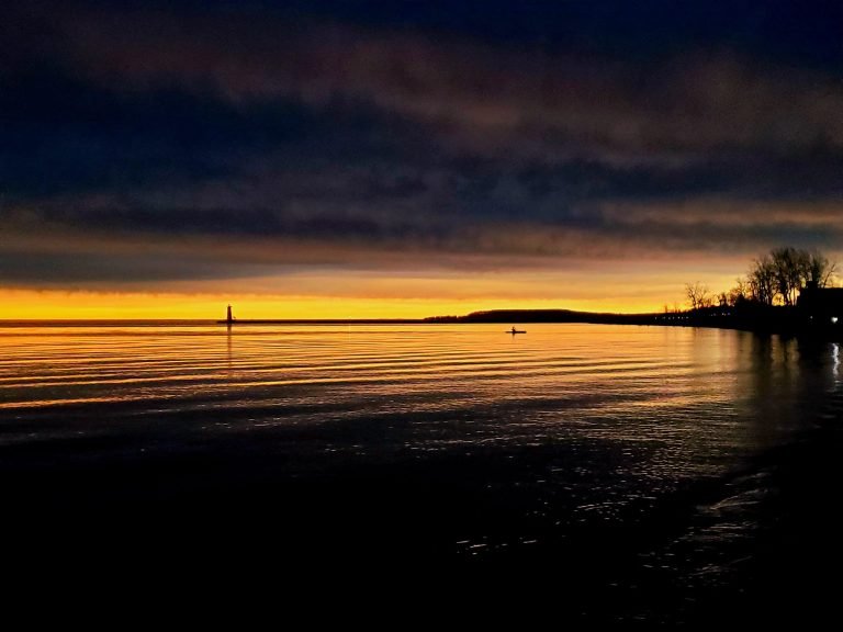 Dark clouds above a golden horizon, a kayaker and a distant pier light silhouetted on the water
