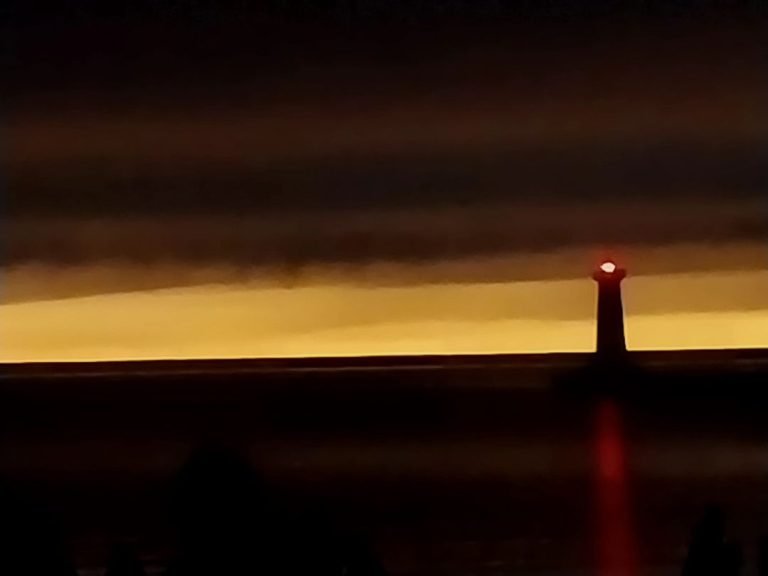 Pier lighthouse silhouetted, its red light glowing under dark clouds and a band of golden sky