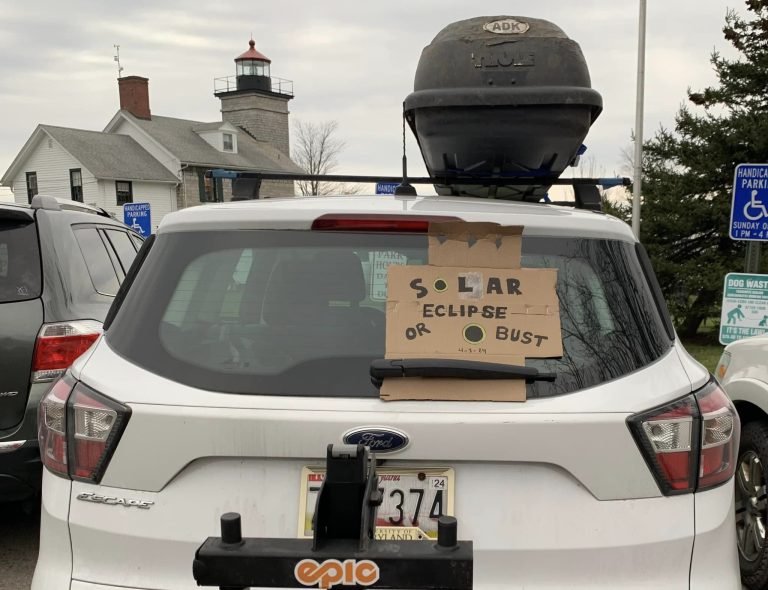Back of a white SUV with a cardboard sign reading 'Solar Eclipse or Bust', a lighthouse behind