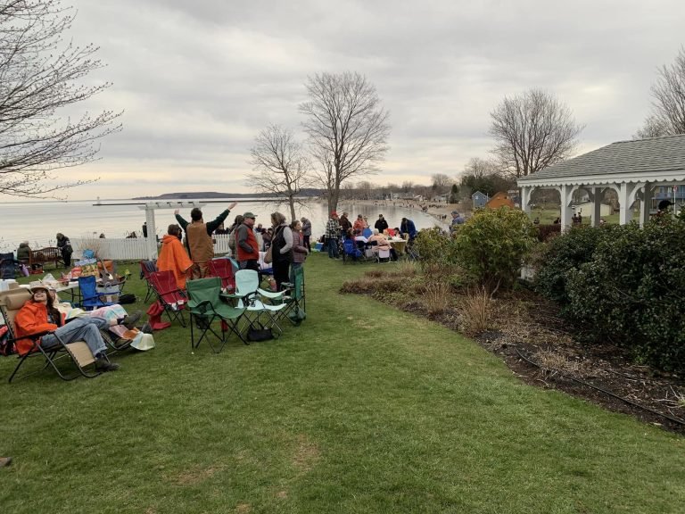Crowd in lawn chairs gathered on a grassy lawn above the bay beside a white gazebo, under an overcast sky