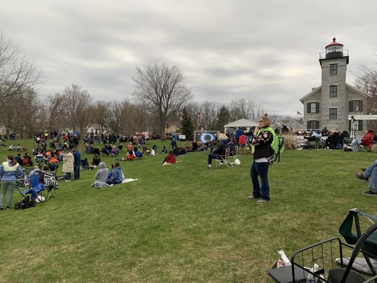 Crowd seated on a lawn near a stone lighthouse, watching a large outdoor screen under a cloudy sky