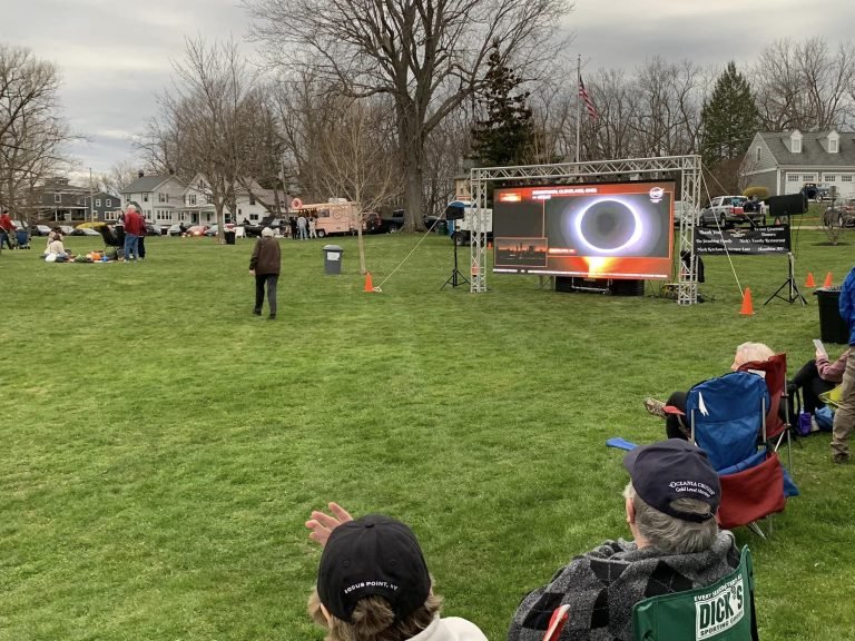 People in lawn chairs in a park watching an outdoor screen that shows the eclipsed sun as a ringed disk