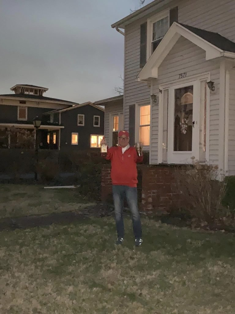 Man in a red jacket and cap raising a bottle and a glass on a darkened front lawn