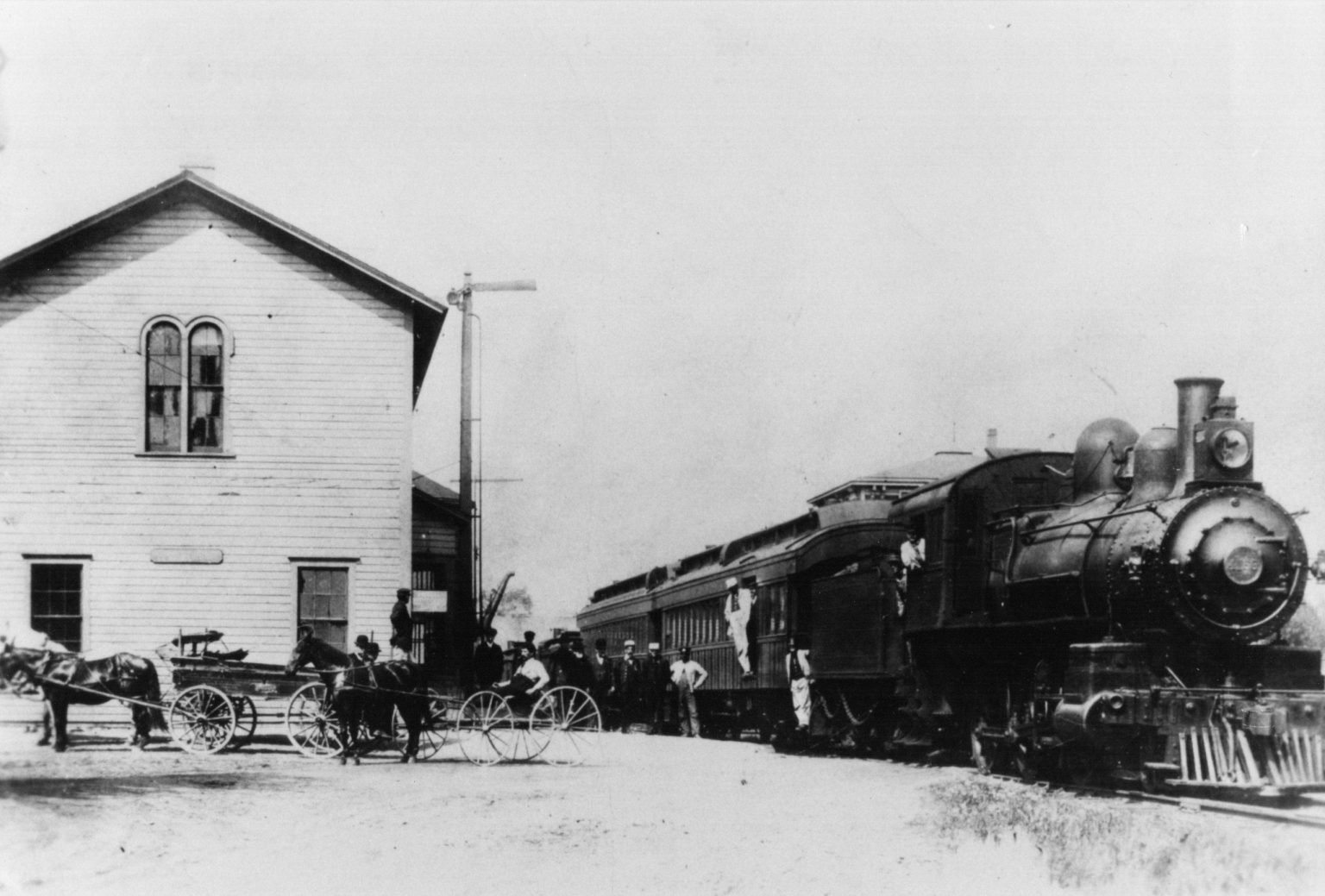 Steam passenger train passing the two-story wooden Bunk House, with horse-drawn wagons and people beside it