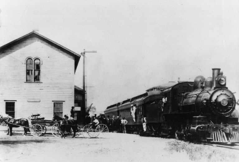 Steam passenger train passing the two-story wooden Bunk House, with horse-drawn wagons and people beside it